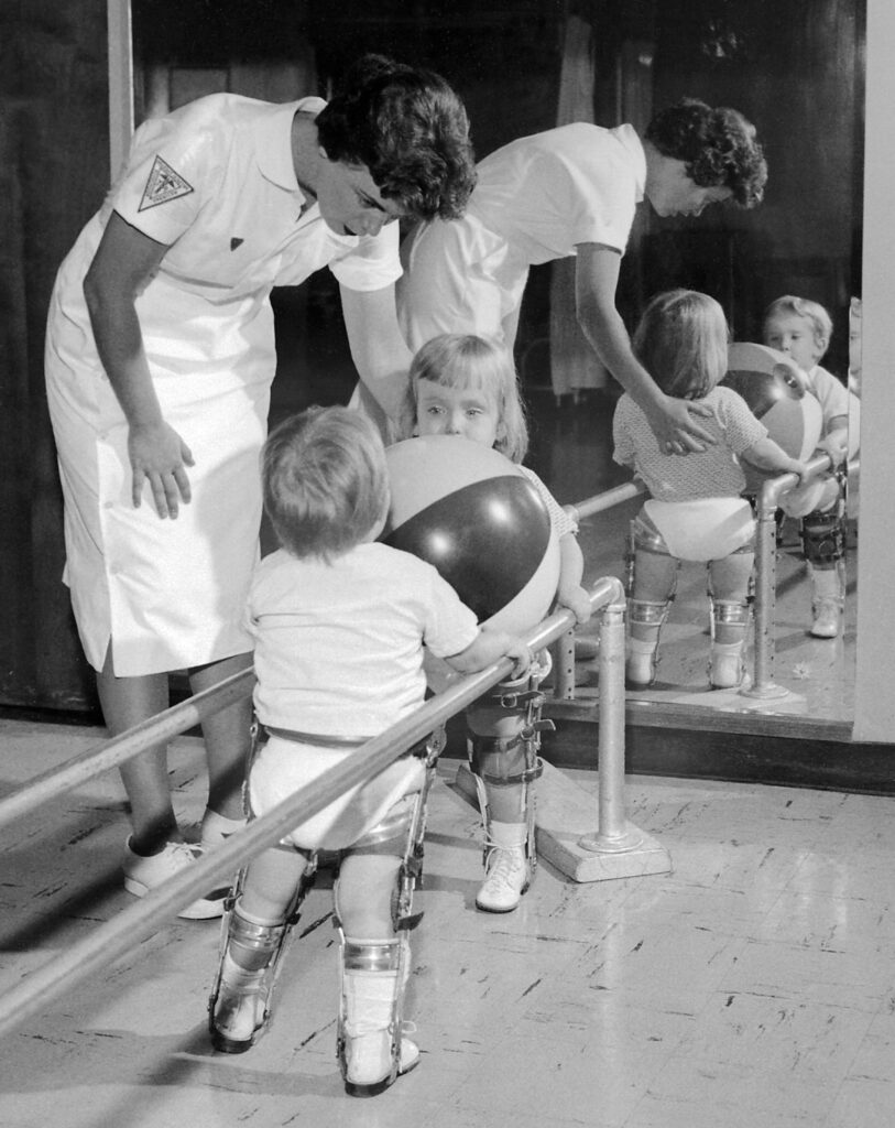 Physical therapist assisting two small children with polio holding on to rail. (Both are in braces). 1963 Charles Farmer